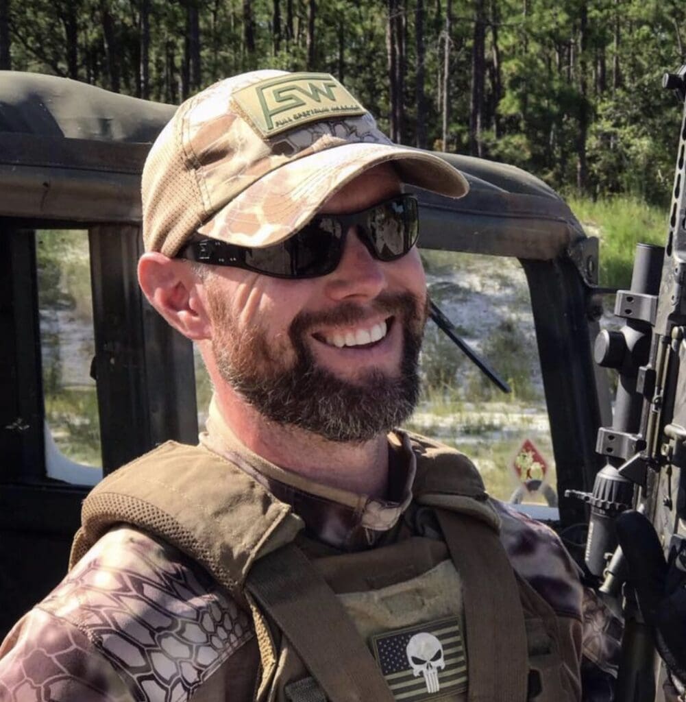A man in camouflage vest and hat standing next to trees.